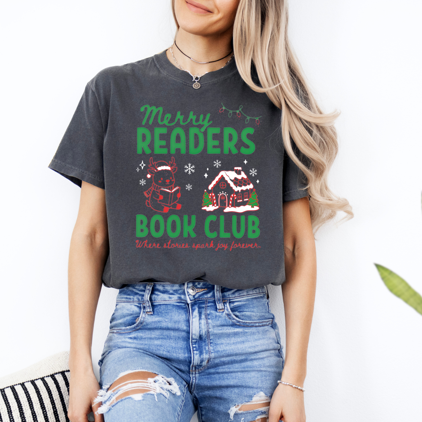 A woman wearing a Pepper Comfort Colors t-shirt that reads 'Merry Readers Book Club' in bold green text, with cute doodle illustrations of a reading reindeer and a a cozy gingerbread house. Underneath is cursive red text that says ' Where stories spark joy forever". Designed by Starlit Prose Bookish Merch Australia.