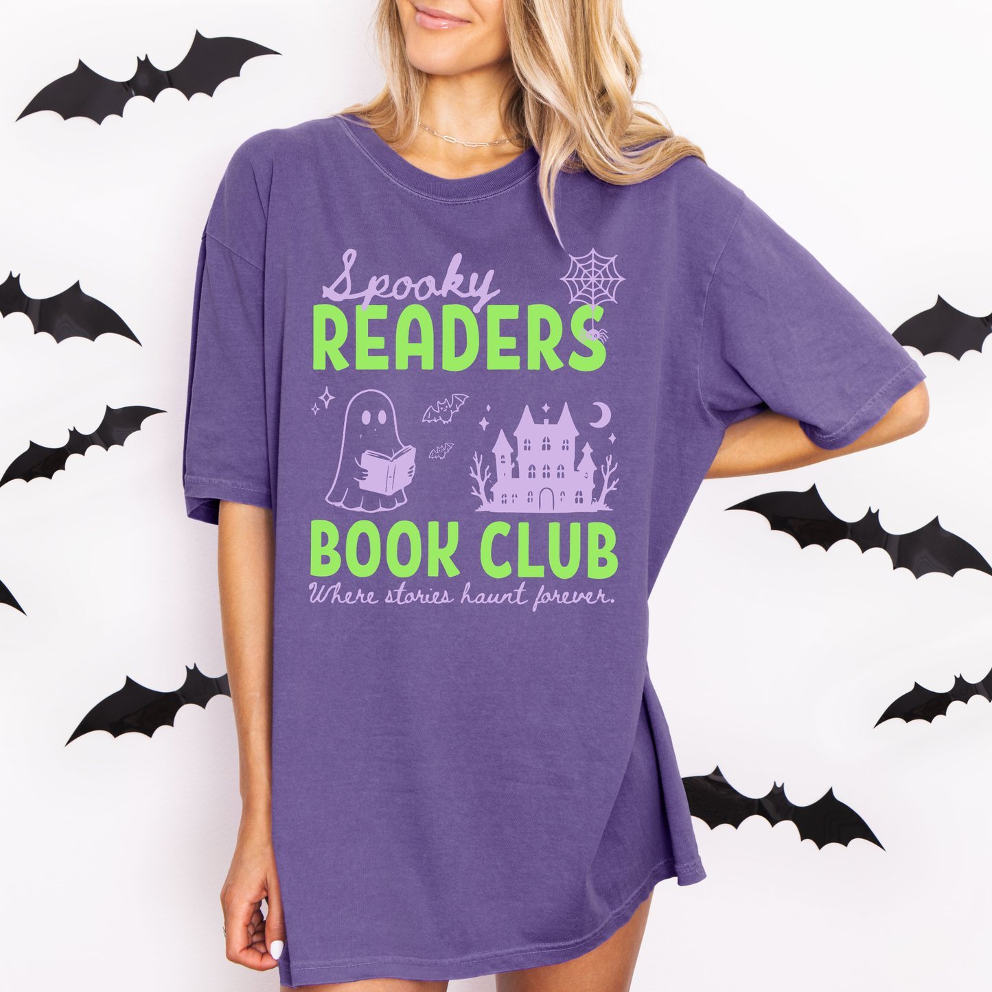  A woman wearing a Grape Comfort colors graphic t-shirt with 'Spooky Readers Book Club' printed in green and purple, with Purple graphics including a cute ghost reading a book, a spider in its web & a haunted mansion paired with distressed blue jeans. Designed by Starlit Prose Bookish Merch Australia