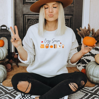 A Woman wearing a white sweatshirt that says "Cozy Season", it includes cute orange Fall Autumn graphics like a stack of books, pumpkin spice latte, pumpkins, a ghost and leaves. Starlit Prose Bookish Merch Australia