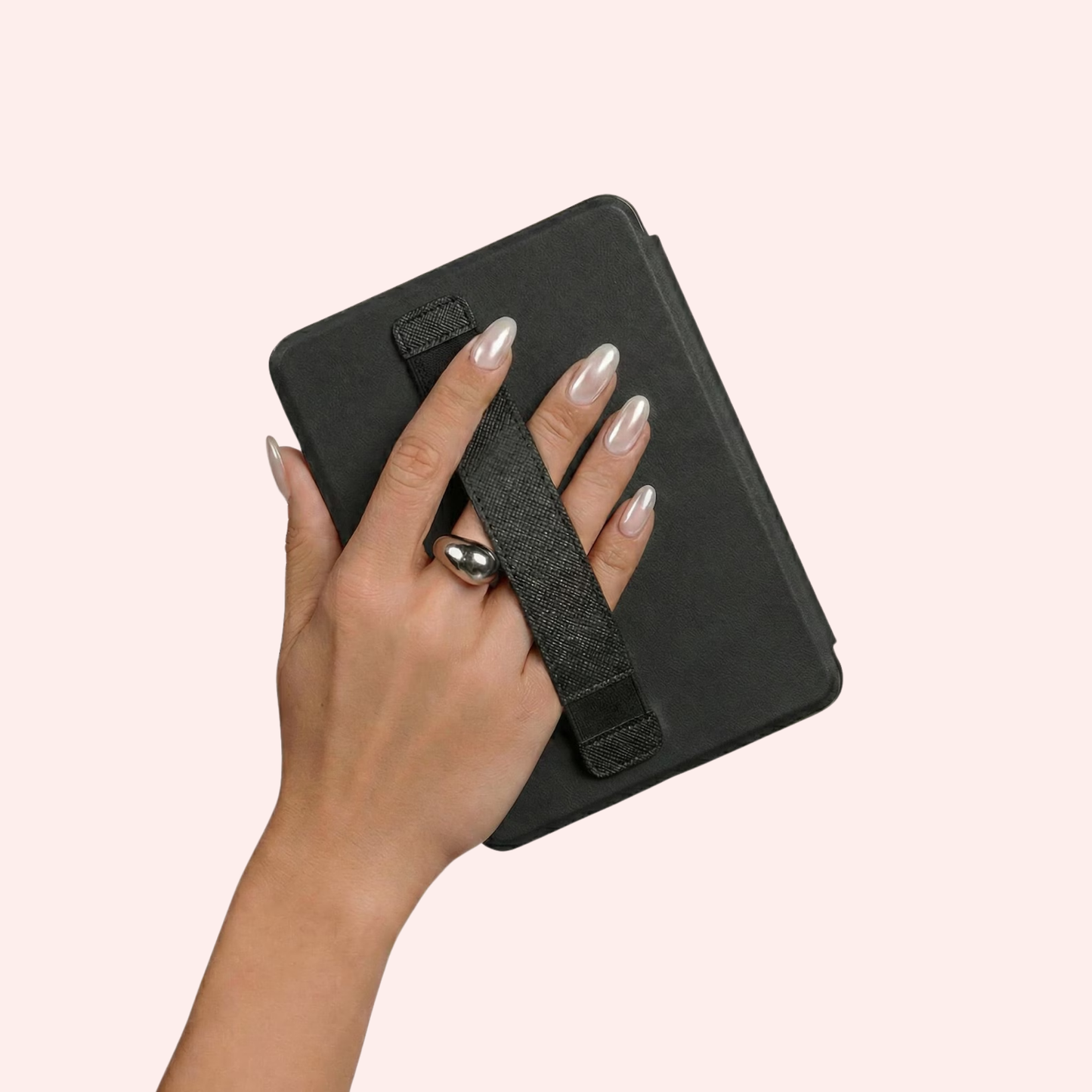 A hand with silver manicured nails holding the kindle case cover, showing the back of the case with a textured black strap, set against a solid pale pink background.