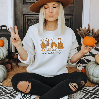 Woman wearing a white sweatshirt with a graphic design of a group of ducks going to a bookstore, sitting in a cozy indoor setting with pumpkins. Starlit Prose Bookish Merch Australia