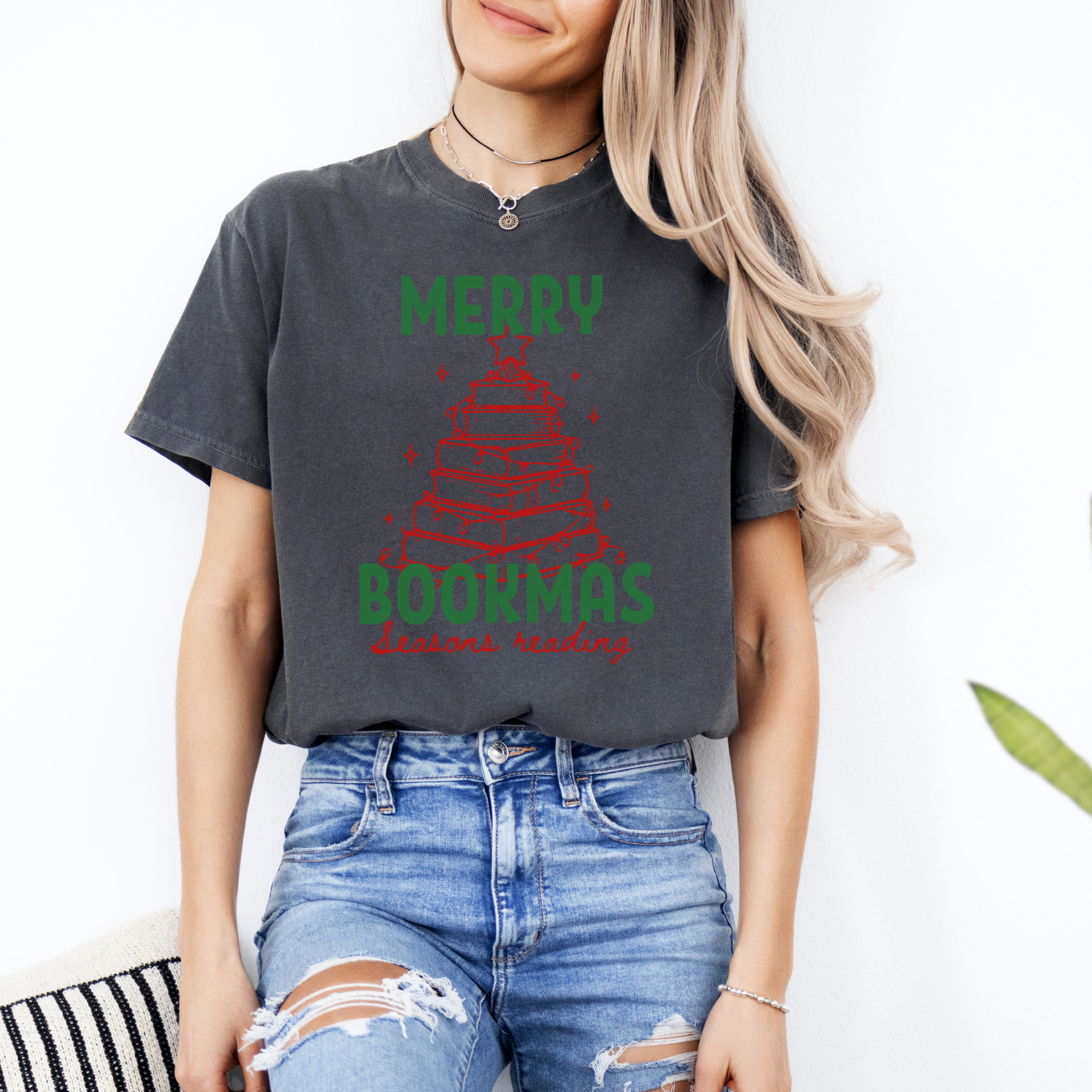 A woman wearing a Pepper Comfort Colors t-shirt with bold text that says "Merry Bookmas" in green, and "Seasons Reading" in red cursive font, includes a doodle illustration of a Christmas tree made of books. Designed by Starlit Prose Bookish Merch Australia.