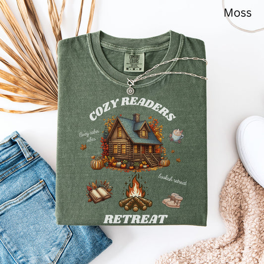 Flay lay of a Moss Green Comfort Colors Tee with 'Cozy Readers Retreat' design including a cozy cabin, pumpkin spice latte, cute slippers, campfire, pumpkins and a book. Starlit Prose Bookish Merch Australia
