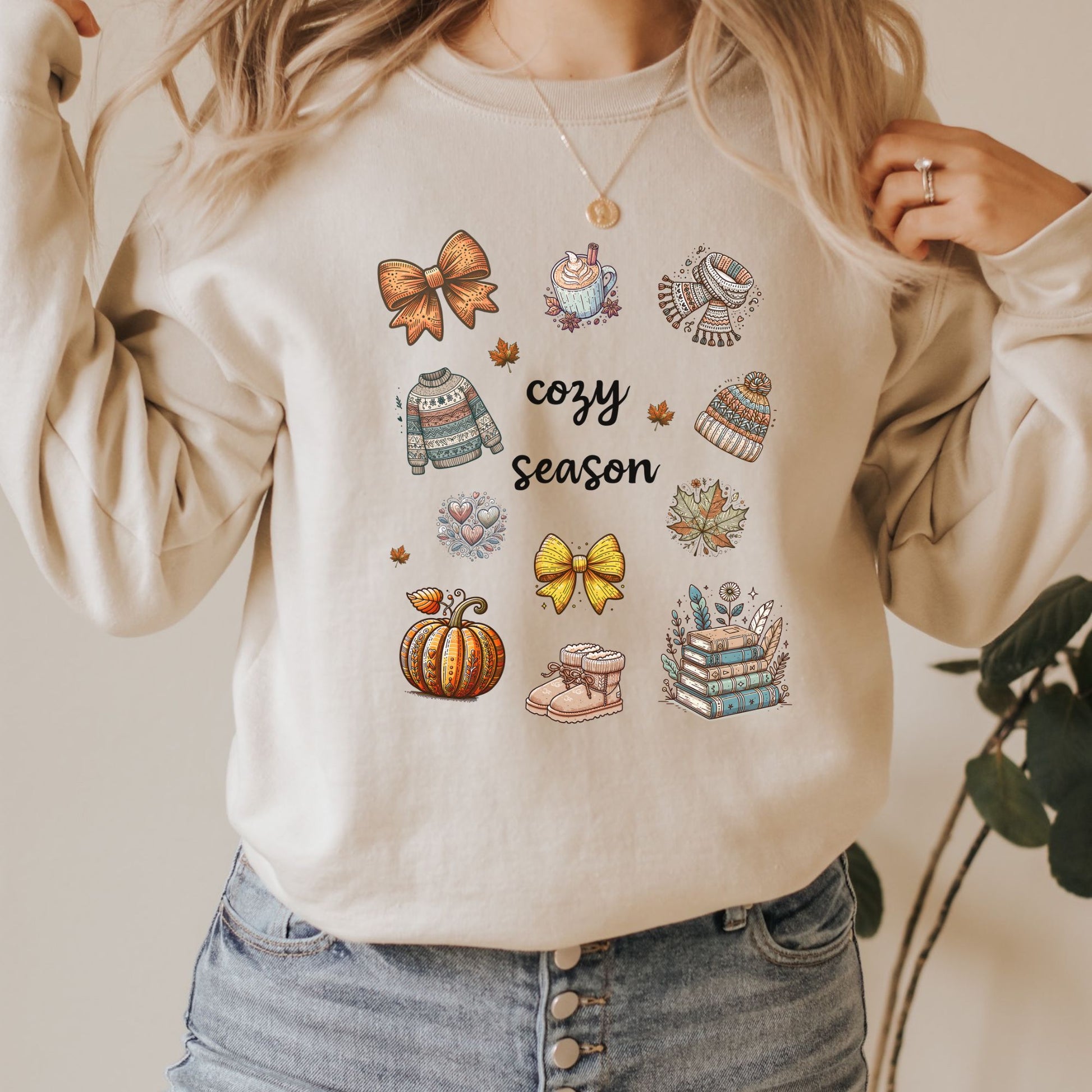 Woman wearing a sand sweatshirt with Fall-themed graphics and text, including a Pumpkin Spice Latte, scarf, beanie, stack of books, sweater & pumpkins sitting in a decorated room with pumpkins. Starlit Prose Bookish Merch Australia