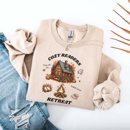 Flat lay of a Sand Sweatshirt with 'Cozy Readers Retreat' design including a cozy cabin, pumpkin spice latte, cute slippers, campfire, pumpkins and a book. Starlit Prose Bookish Merch Australia