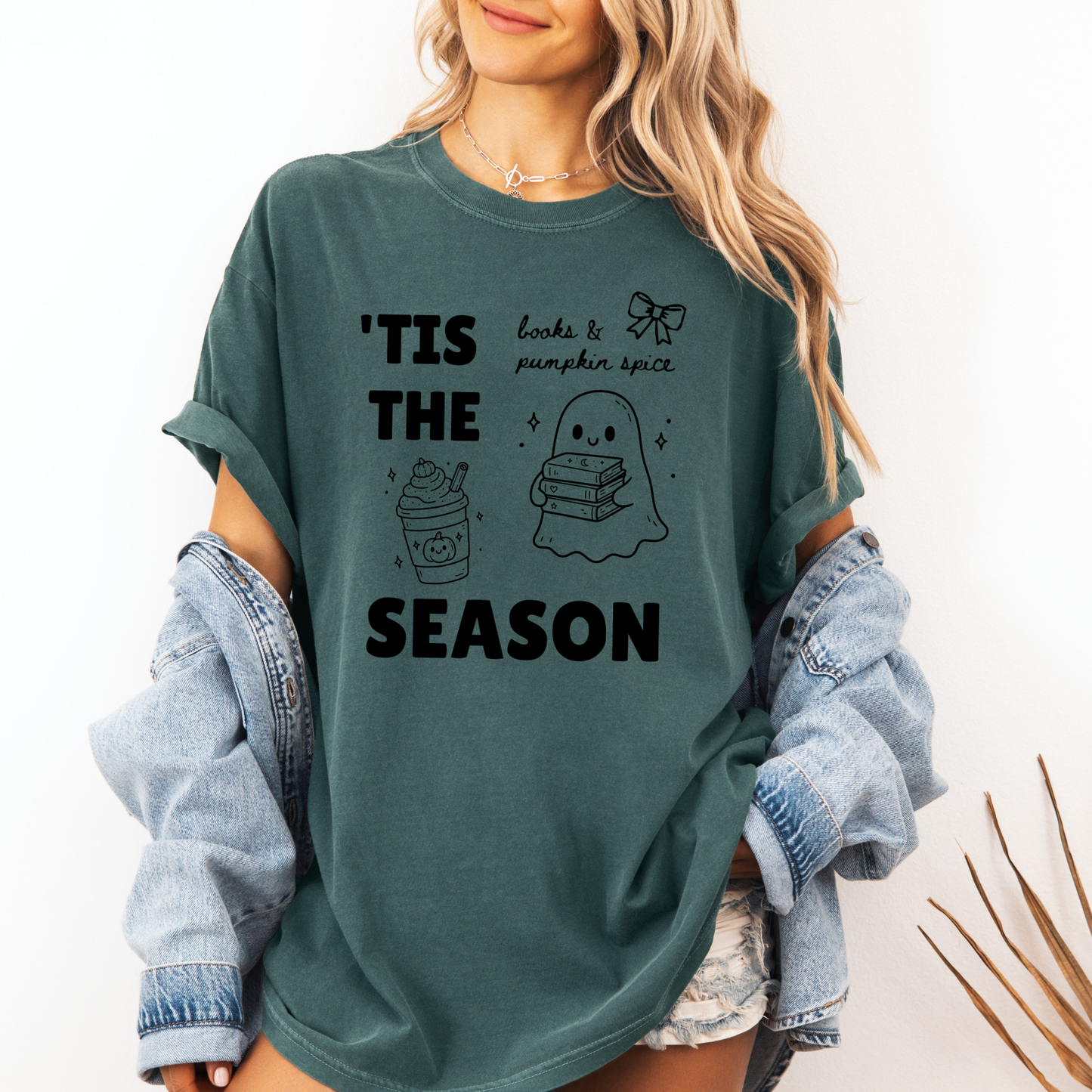 A woman wearing a Seafoam Green graphic t-shirt with 'TIS THE SEASON' in bold font with cute Halloween-themed illustrations including a ghost holding a stack of books, a cute pumpkin spiced latte and a coquette bow with the phrase "books & pumpkin spice". Designed by Starlit Prose Bookish Merch Australia.