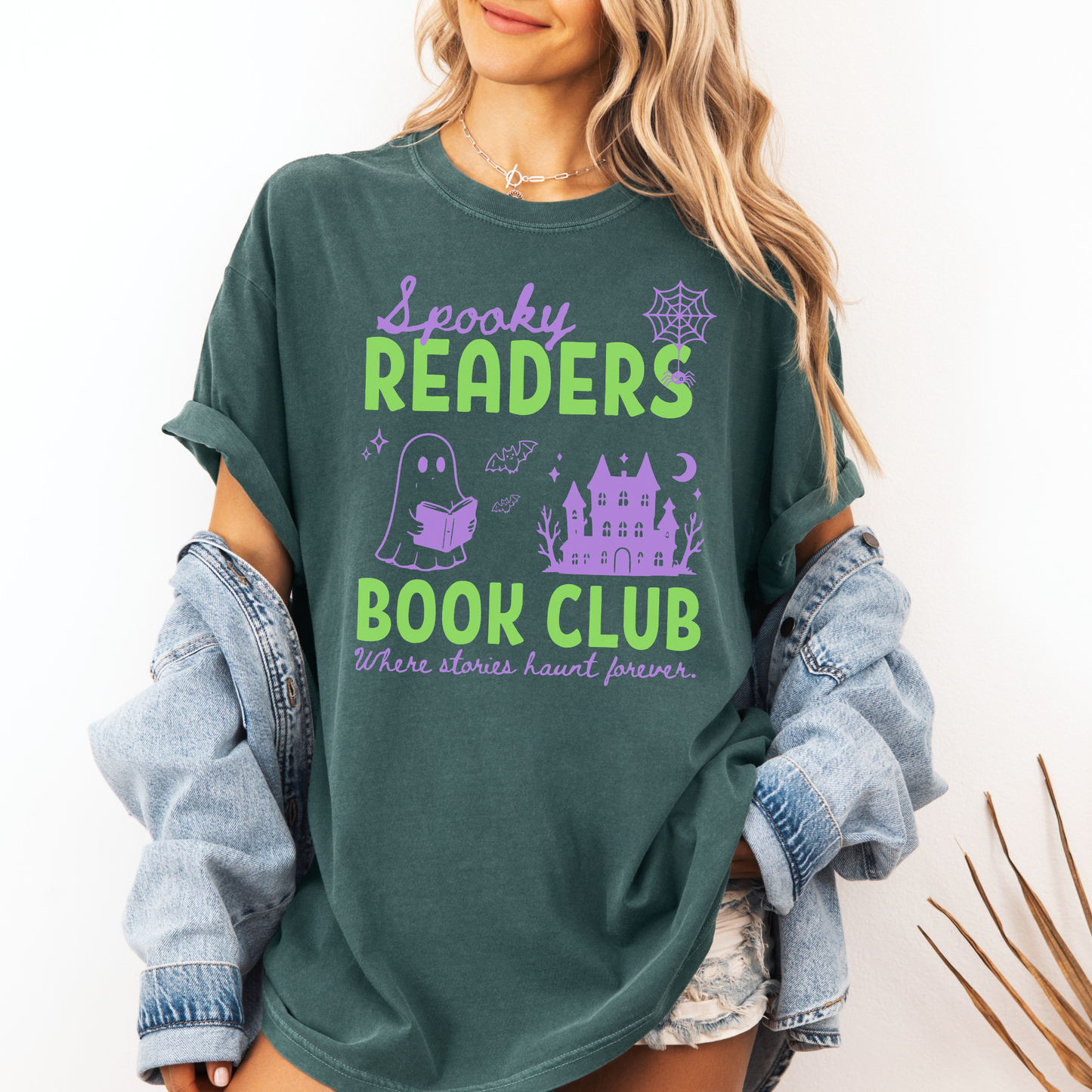  A woman wearing a Seafoam green Comfort colors graphic t-shirt with 'Spooky Readers Book Club' printed in green and purple, with Purple graphics including a cute ghost reading a book, a spider in its web & a haunted mansion paired with distressed blue jeans. Designed by Starlit Prose Bookish Merch Australia