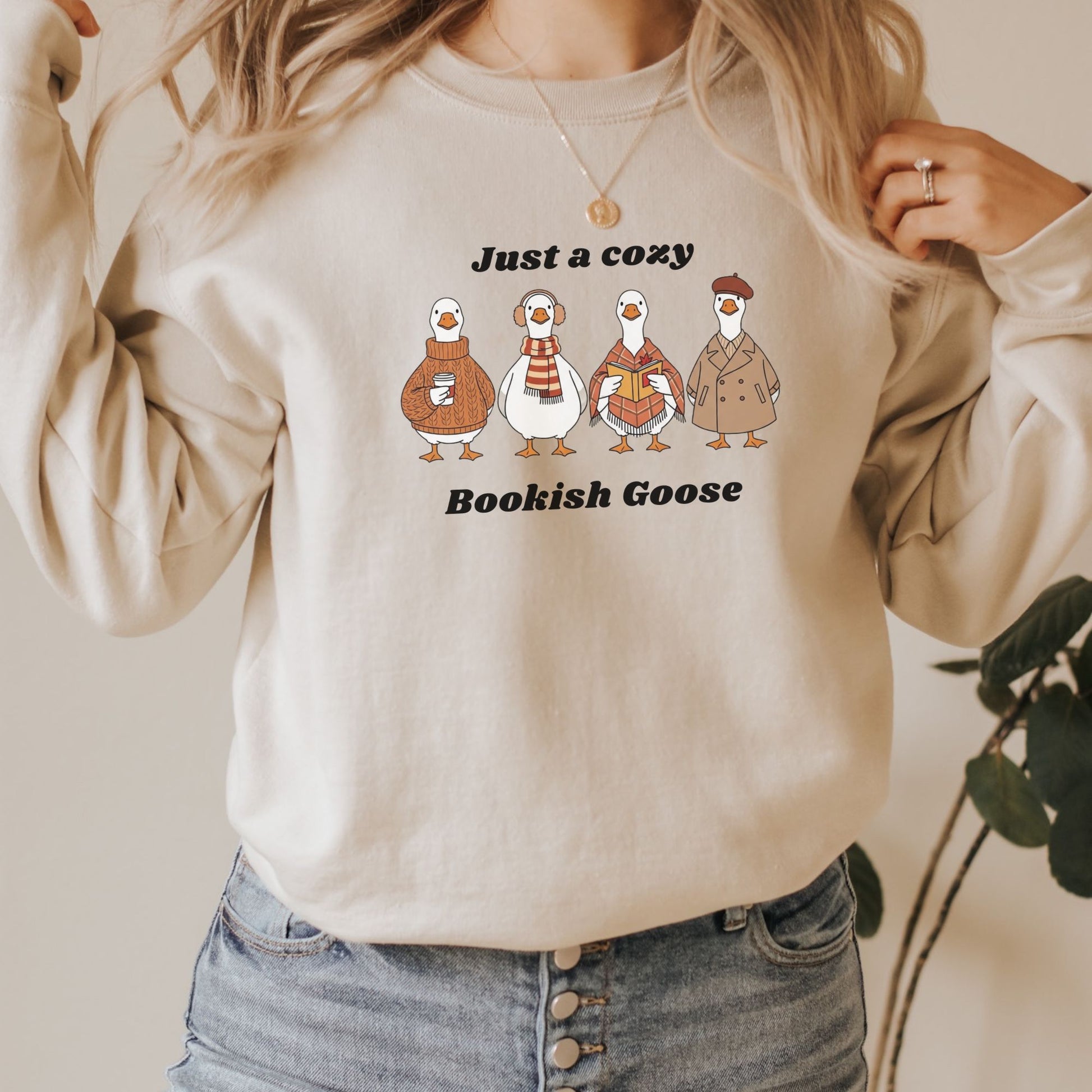 Sand colour sweatshirt with cartoon geese and text  that says "Just a Cozy Bookish Goose" worn by a person, with a neutral background. Starlit Prose Bookish Merch Australia