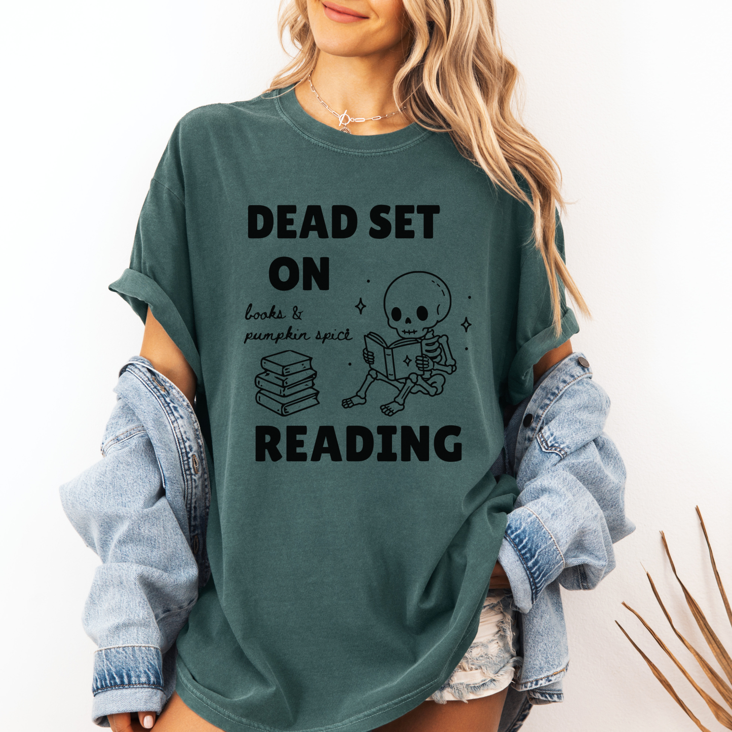 A woman wearing a Seafoam Green Comfort Colors  t-shirt with a black print that reads 'DEAD SET ON READING' alongside a skeleton reading books and a stack of books. It also says books & pumpkin spice in script font. Designed by Starlit Prose Bookish Merch Australia