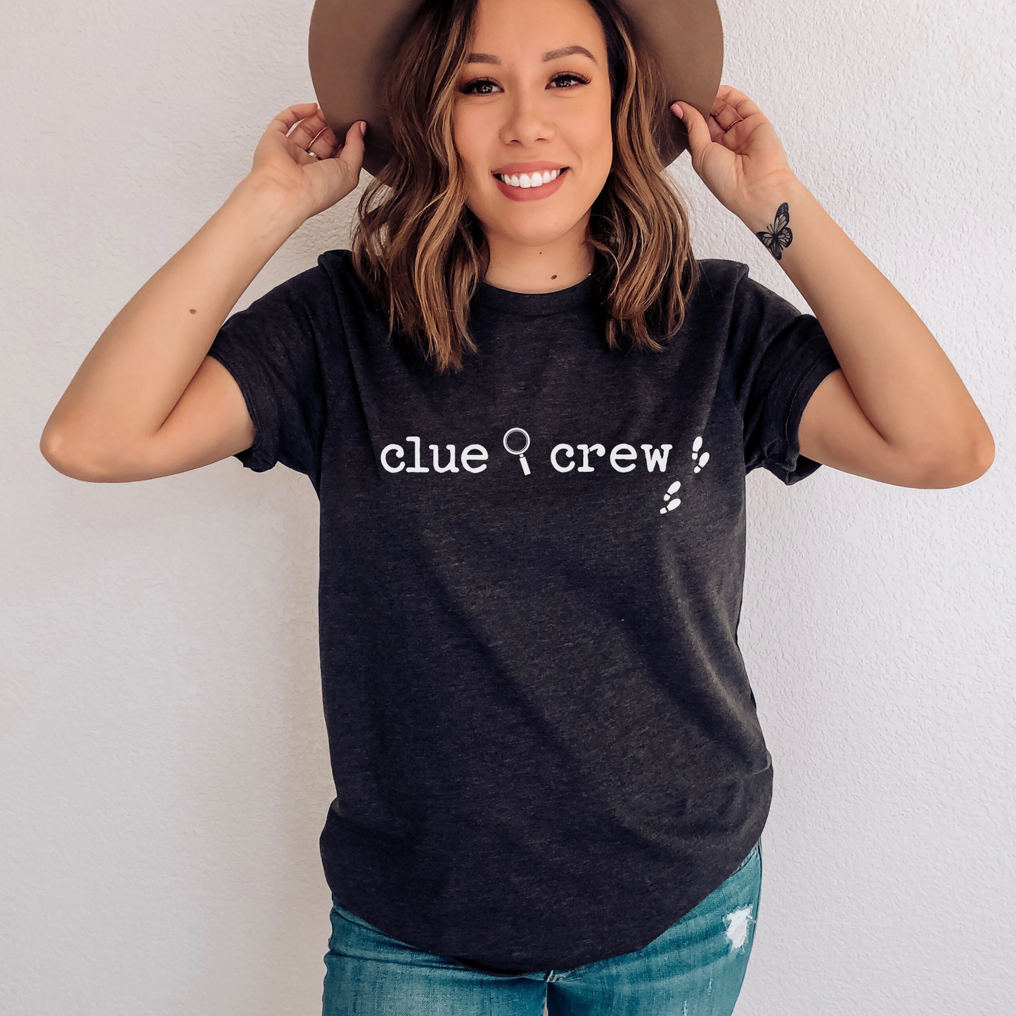 Woman wearing a Dark grey heather t-shirt with 'clue crew' text, holding a brown hat.