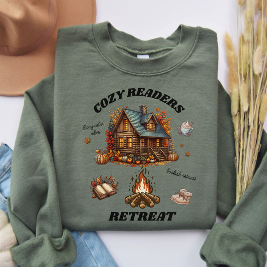 Flat lay of a Green Sweatshirt with 'Cozy Readers Retreat' design including a cozy cabin, pumpkin spice latte, cute slippers, campfire, pumpkins and a book. Starlit Prose Bookish Merch Australia
