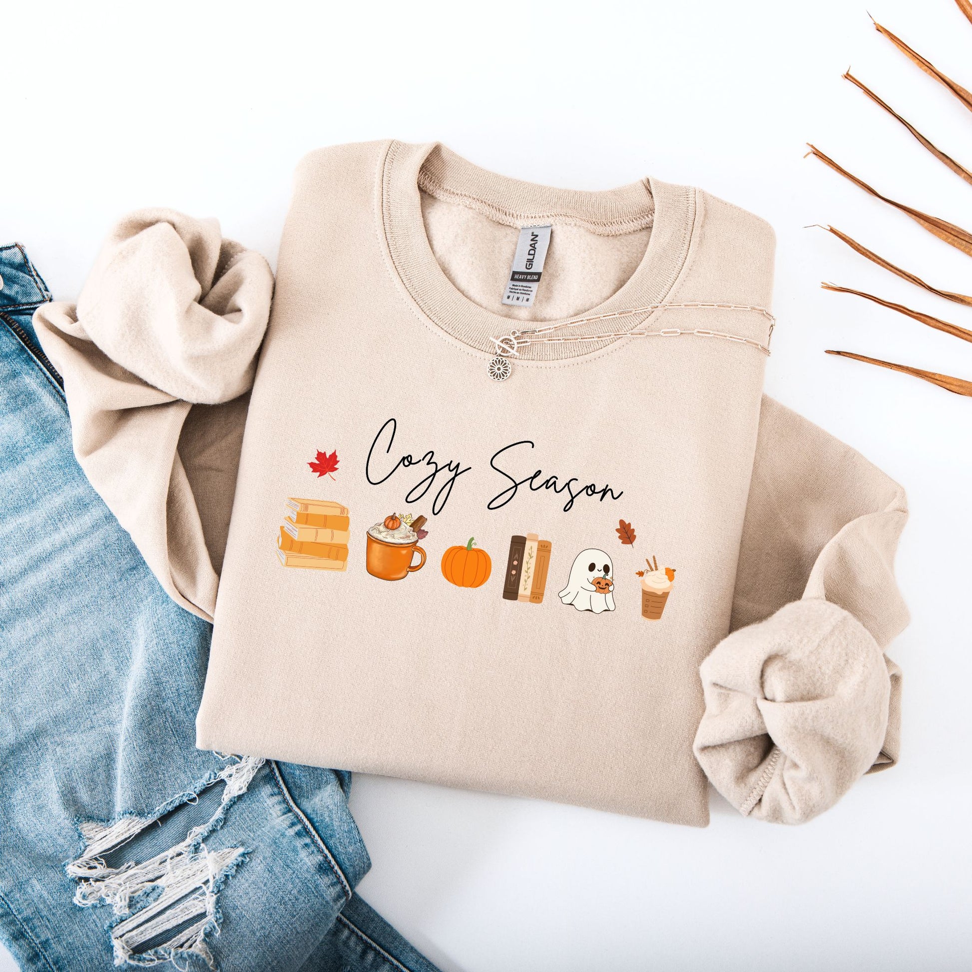 Flatlay of  a Sand sweatshirt that says "Cozy Season", it includes cute orange Fall Autumn graphics like a stack of books, pumpkin spice latte, pumpkins, a ghost and leaves. Starlit Prose Bookish Merch Australia