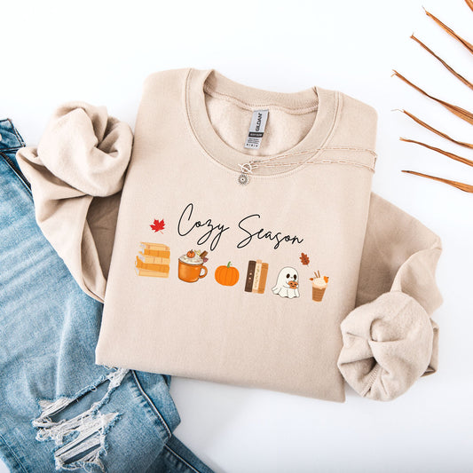 Flatlay of  a Sand sweatshirt that says "Cozy Season", it includes cute orange Fall Autumn graphics like a stack of books, pumpkin spice latte, pumpkins, a ghost and leaves. Starlit Prose Bookish Merch Australia