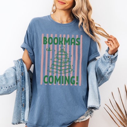 A woman wearing a Blue Jean Comfort Colors t-shirt, it has a pink distressed stripe pattern in the background, with bold green lettering that says "Bookmas is Coming!", it includes a green doodle illustration of a Christmas Tree made of books. Designed by Starlit Prose Bookish Merch Australia.