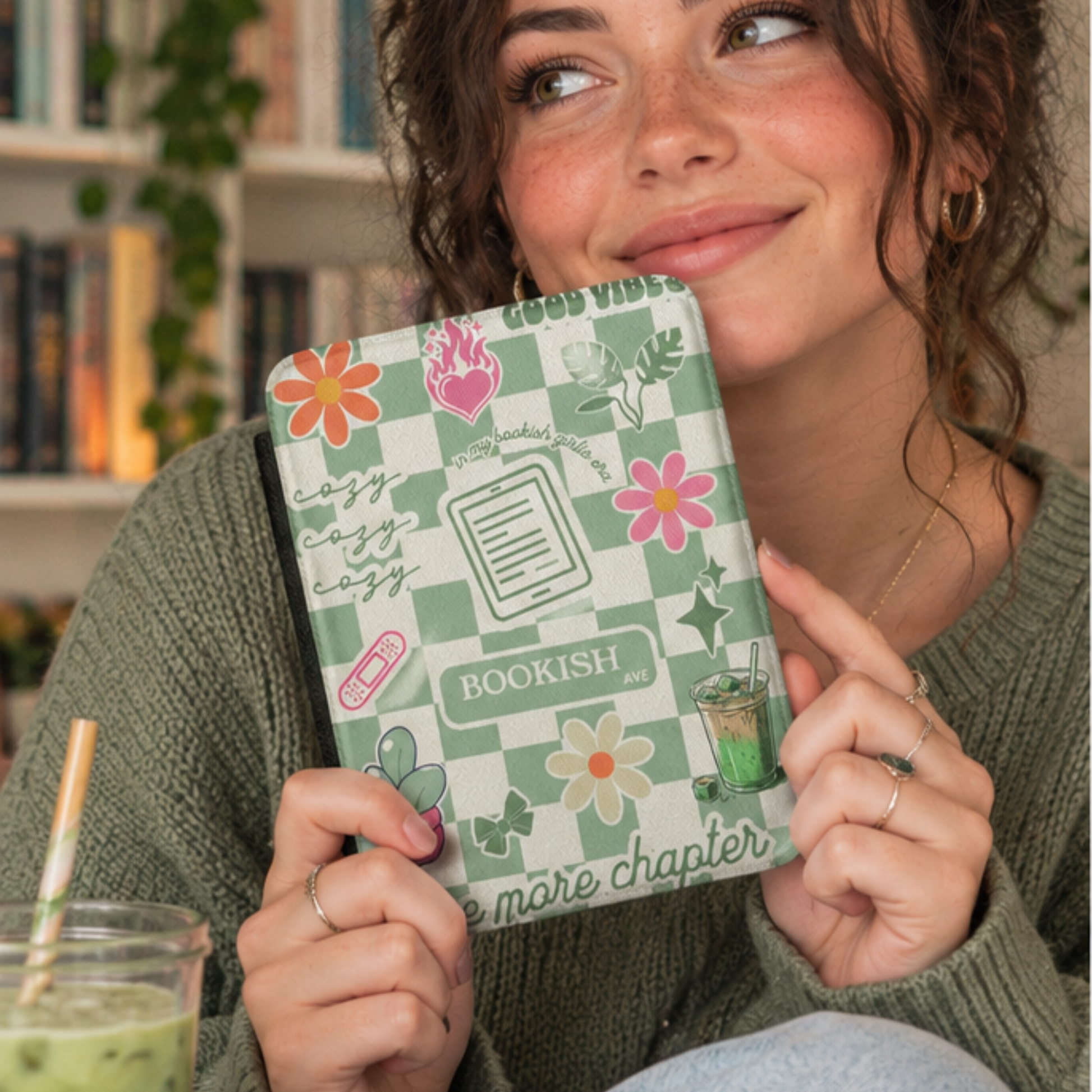A woman in a green knit sweater holds a Matcha Green Bookish Kindle Case in front of her face. The case has a Matcha green and white checkerboard pattern with floral illustrations, the words 'BOOKISH AVE' and 'GOOD VIBES', One more Chapter, cozy cozy cozy and includes small decorative illustrations like matcha iced latte, coquette bows and a kindle. Designed by Starlit Prose Bookish Merch Australia.