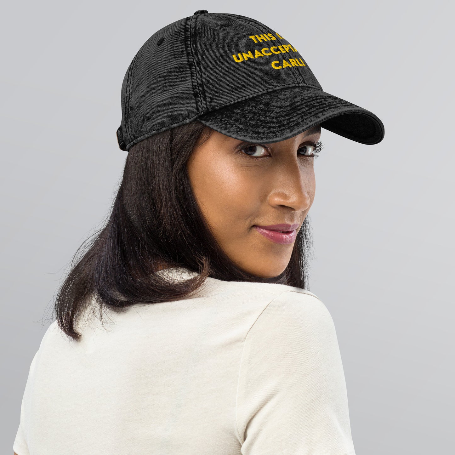 Woman wearing A washed black denim baseball cap that says "This is unacceptable Carl" in yellow embroidered text. Quote from Princess Donut of Dungeon Crawler Carl book series. Designed by Starlit Prose Bookish Merch Australia